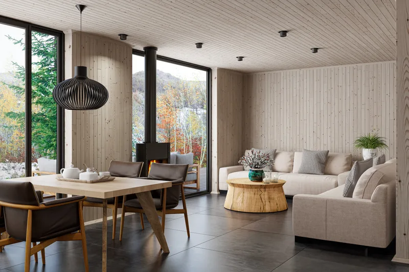 Open-plan dining room with a pale wood table and four armchairs by tall windows, a wood stove and a cream sofa beyond