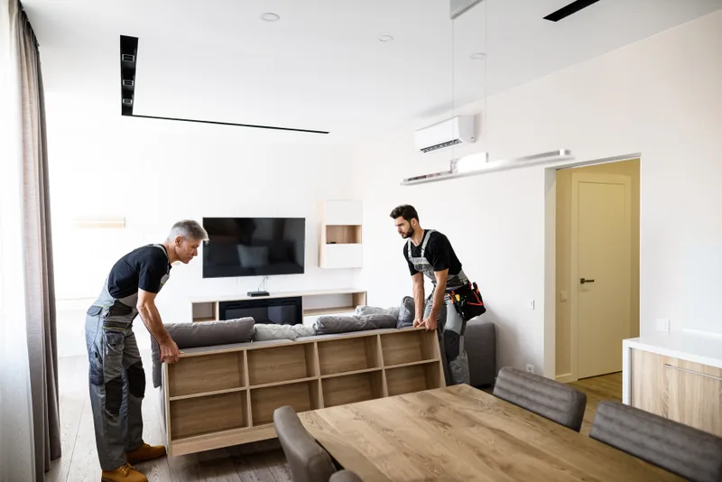 Two delivery crew members in work aprons lifting a low wooden shelving unit into place in a bright living room