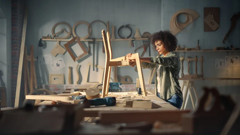 A furniture maker checking the joints of a bare wooden chair frame at a workbench, templates and tools hanging on the wall behind her