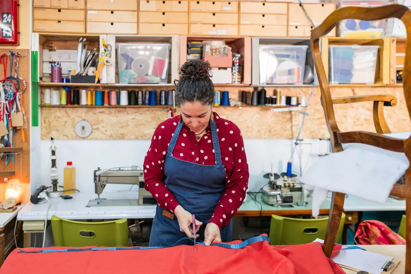 An upholsterer in a denim apron measuring and cutting red fabric on a cutting table, spools of thread and a chair frame around her