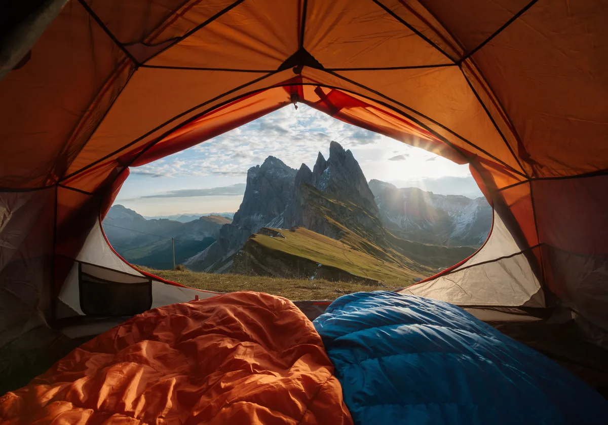 Dawn light on jagged peaks seen through the open door of an orange tent, two sleeping bags in the foreground