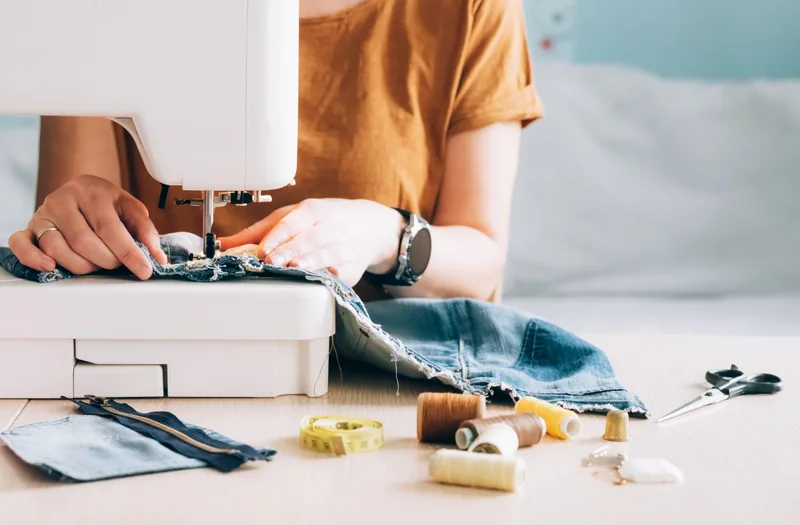 Repair tech feeding a worn fabric panel through a sewing machine, spools of thread and a cut patch on the bench