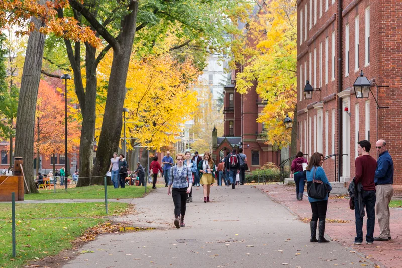 Students walking along a tree-lined brick path between red-brick university halls in full autumn color