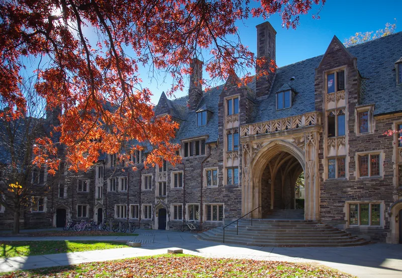 Stone collegiate-gothic residence hall with an arched entryway and slate roofs, framed by red autumn leaves, with bicycles parked outside