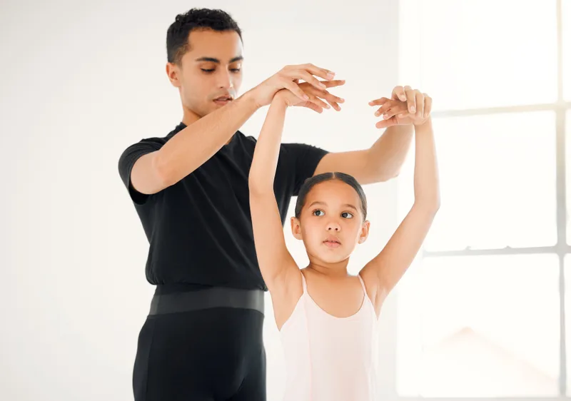 A ballet teacher lifting a young student's arms into position in a bright studio