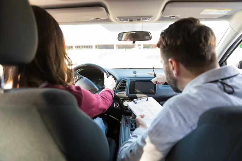 Instructor in the passenger seat pointing at the dashboard while an adult student drives