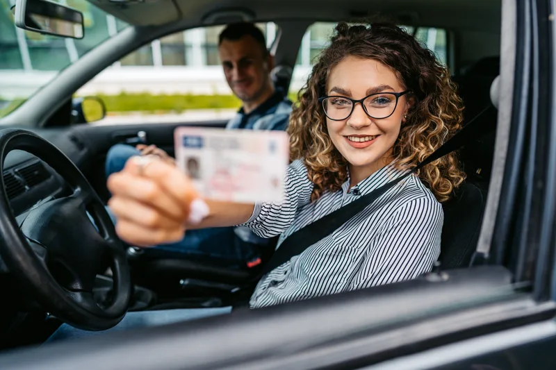 Young driver in glasses holding up her brand-new license from the driver's seat, grinning