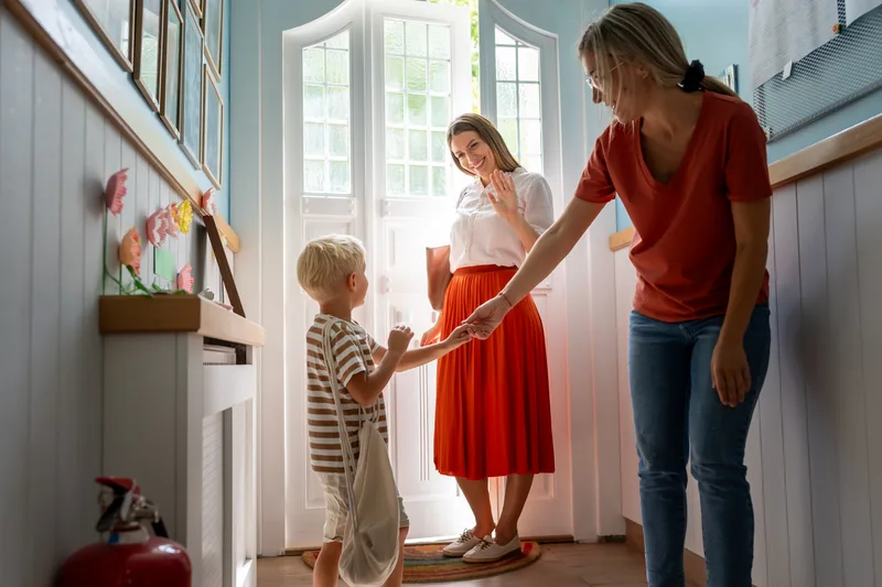 A teacher greets a parent and a young child at the classroom door in the morning