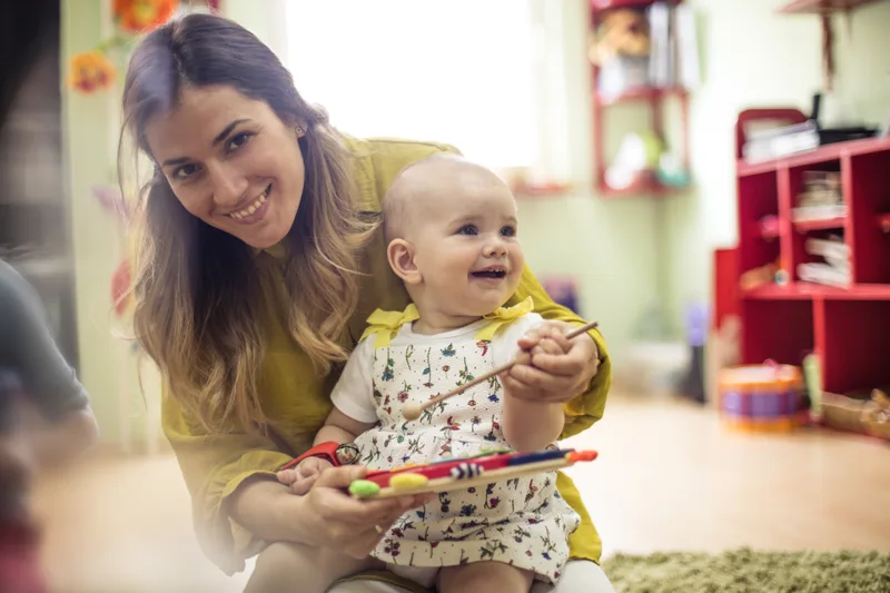 A caregiver smiles while holding a baby on her lap in a bright infant room