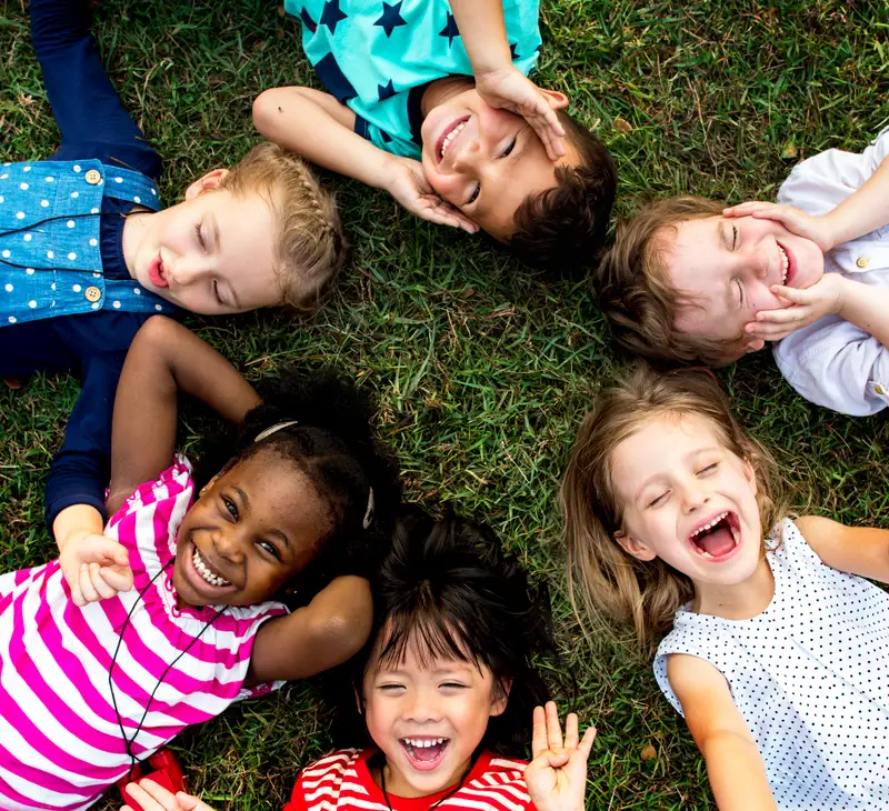 A group of children lie on the grass in a circle, laughing and looking up