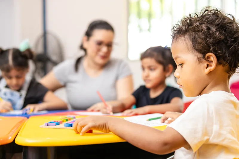 Pre-K children practise letters with a teacher at a bright yellow table