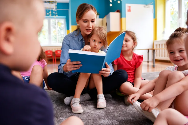 A teacher reads a picture book to preschoolers sitting together on a classroom rug