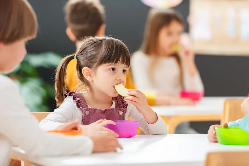 A young child eats a snack at a classroom table with friends