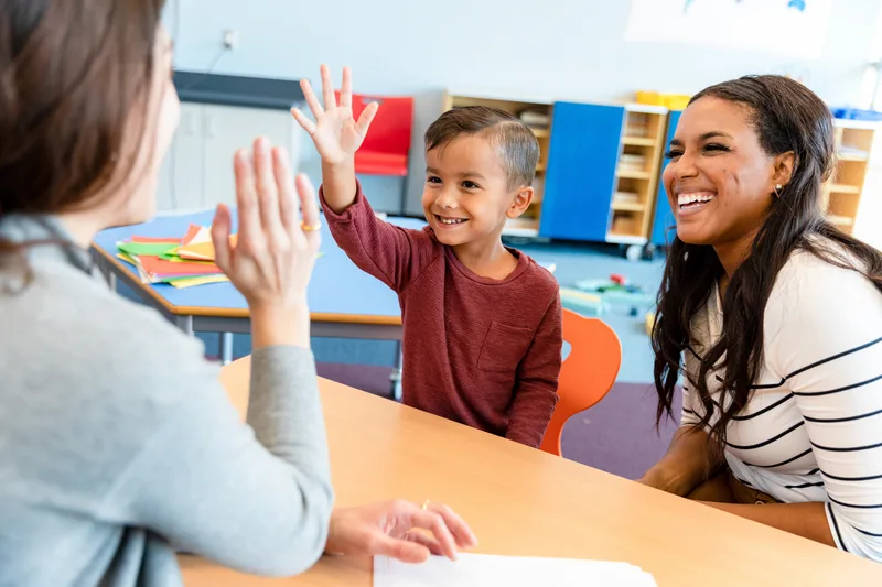 A smiling teacher gives a preschool boy a high five at a classroom table