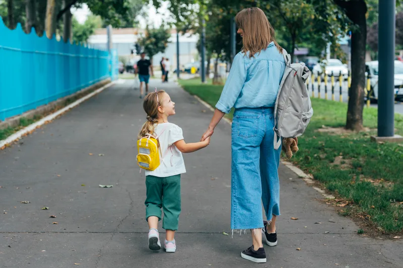 A parent and a child with a yellow backpack walk hand in hand along a school path