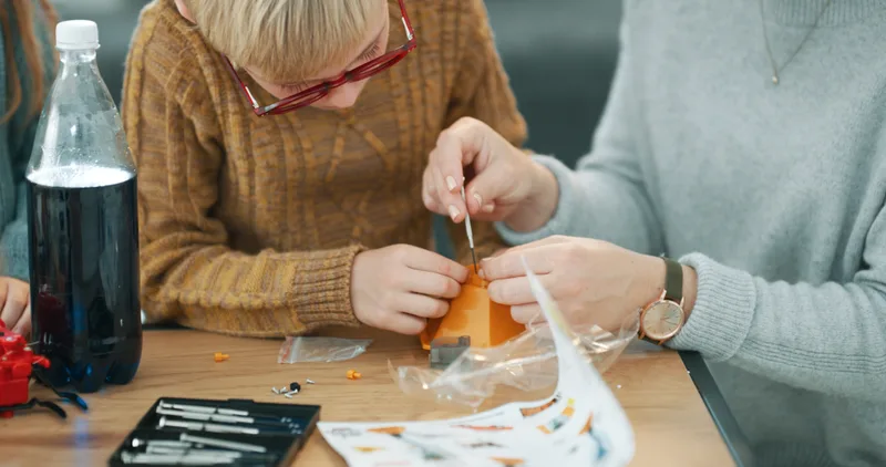 An adult's hands guiding a student's hands as they cut and assemble a small science project at a table