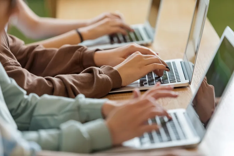 Close-up of several teenagers' hands typing on laptops side by side at a long table