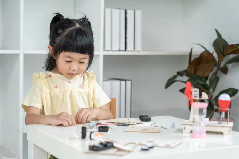 A young student at a white table connecting wires and small parts on a wooden circuit kit