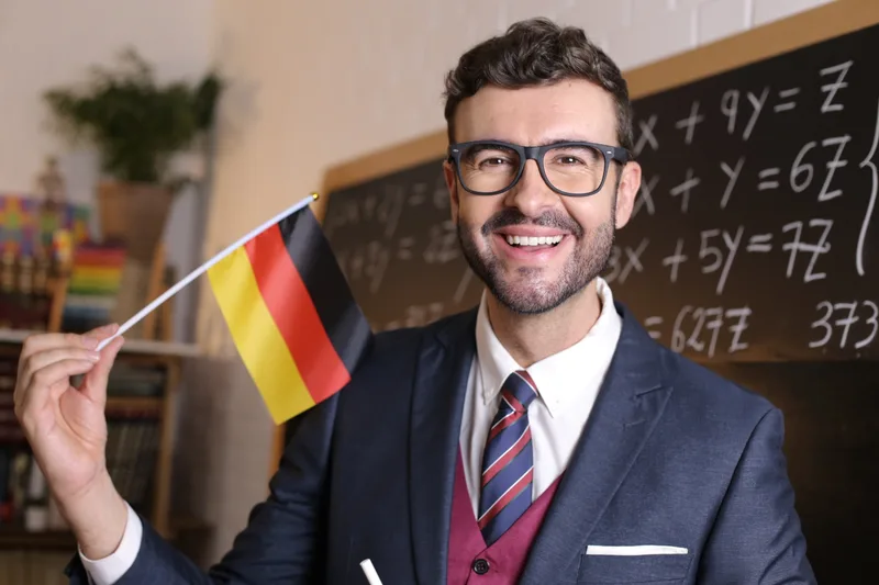 A smiling world-languages teacher in glasses and a suit holds up a small German flag in front of a chalkboard covered in equations