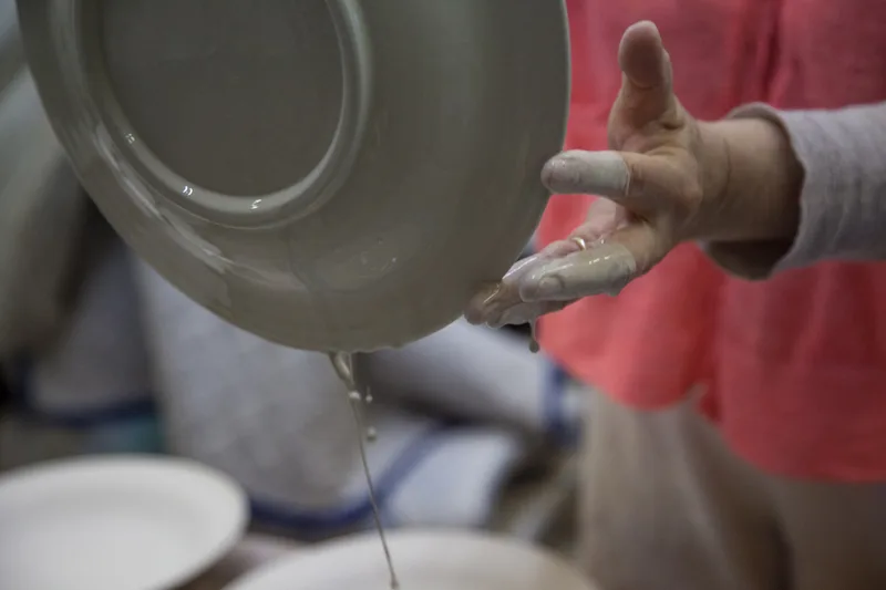A potter's hand lifting a plate out of a bucket of glaze, with glaze dripping from the rim
