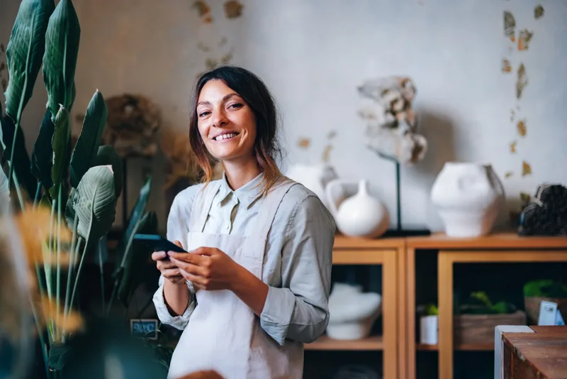 Marisol Vance in an apron, smiling with her phone in a shop of white ceramic vases