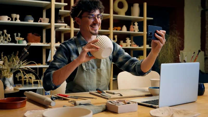 Nico Ferrante in an apron in his studio, filming himself on his phone while holding up a ribbed white bowl, with shelves of pots behind him