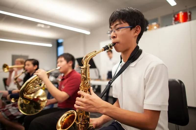 Teenage student playing alto saxophone in a band classroom with other young musicians behind him