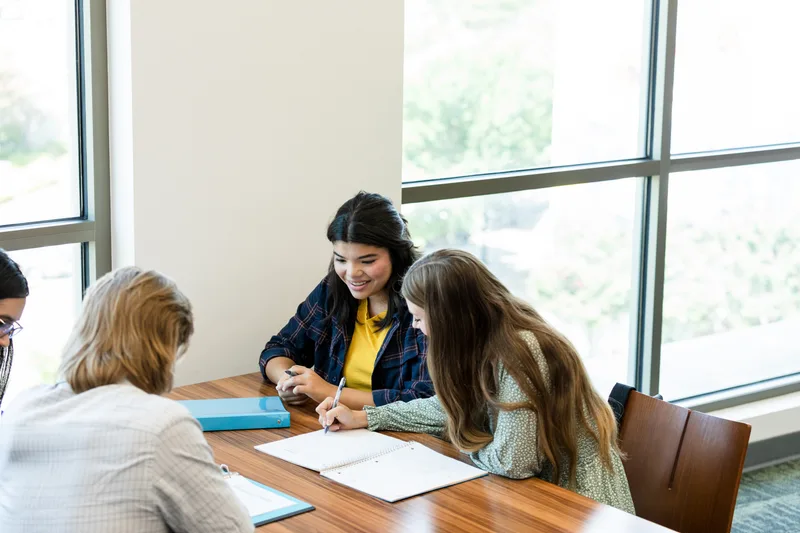 Students working together at a shared table by tall windows, one writing in a spiral notebook