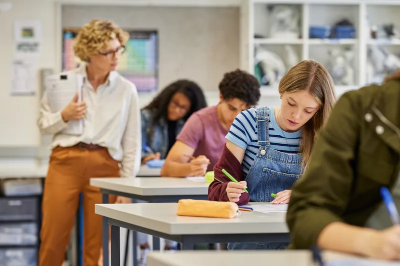 High-school students writing a timed practice exam at individual desks while a proctor walks the room