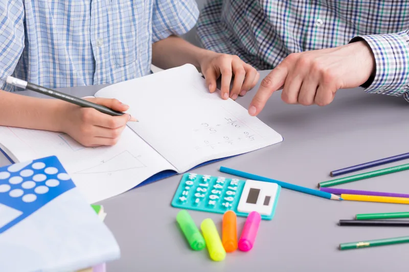 A tutor points to a step in an elementary student's math notebook, highlighters and a calculator on the table