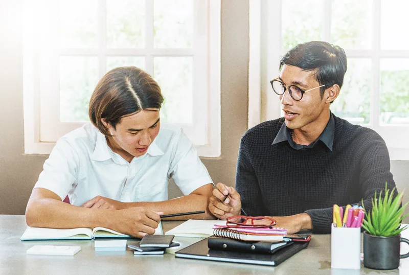 A tutor and a teenage student working through a problem together at a bright table by the window