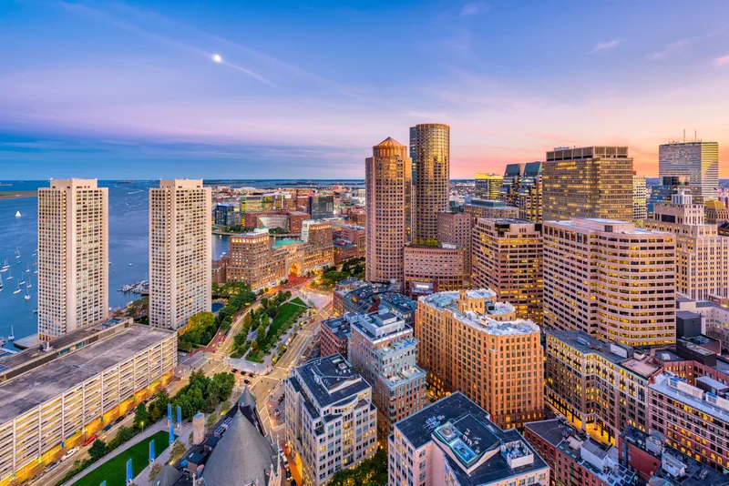 Aerial view of Boston's financial district and harbor at dusk, sailboats moored along the waterfront