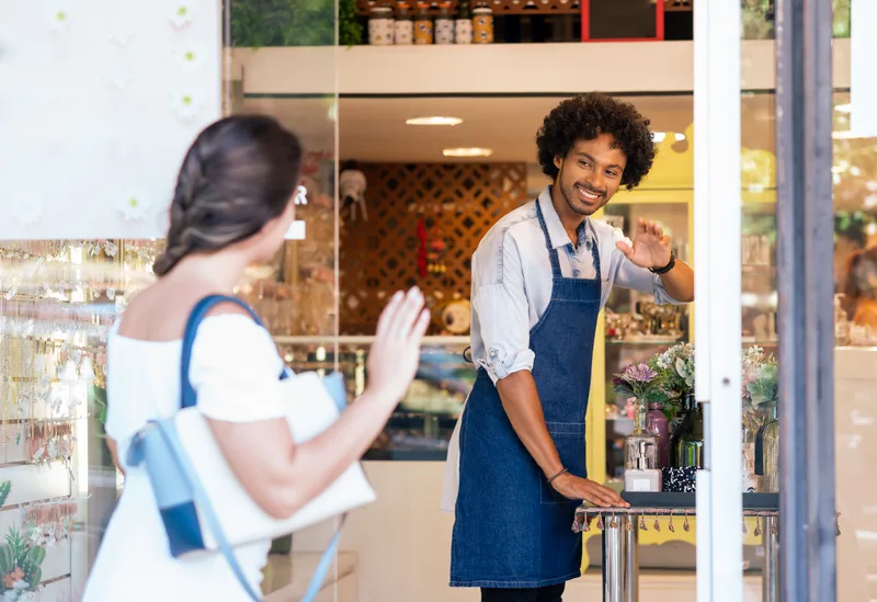 A boutique owner in a denim apron waving hello to a customer walking through the shop door