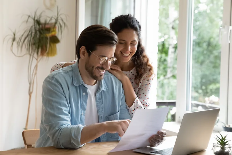 A couple at a kitchen table reading a letter together and smiling, a laptop open beside them