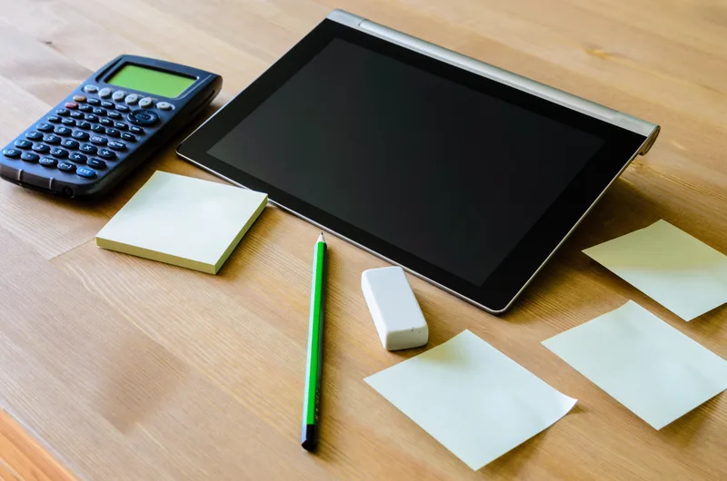 A tidy desk from above with a calculator, a tablet, a green pencil and blank sticky notes