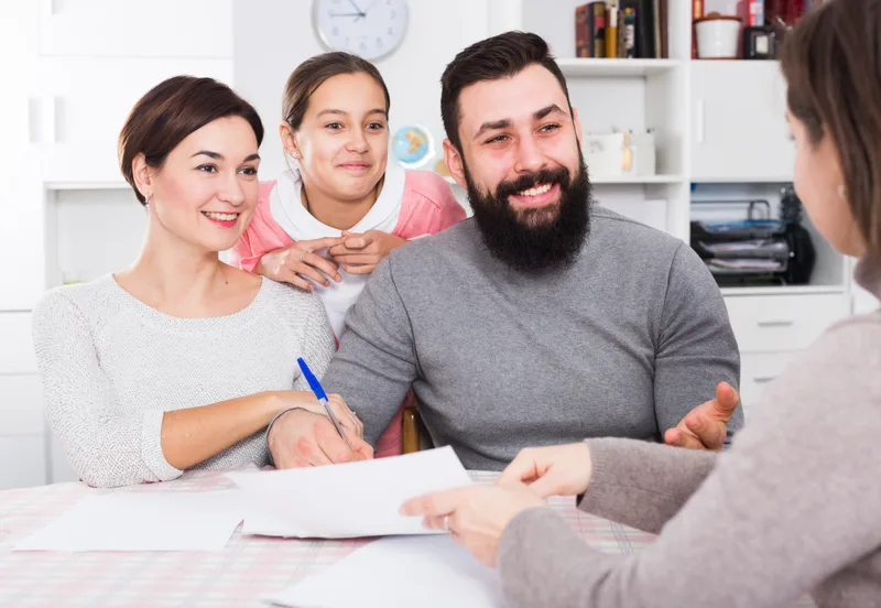 A smiling couple and their daughter at a desk, the mother signing a form as the preparer across the table points to the signature line
