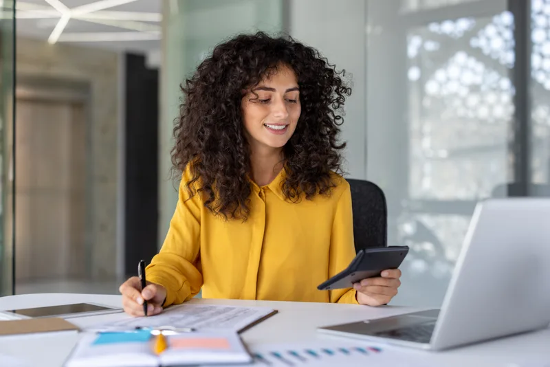 A preparer in a yellow blouse working at a bright office desk with a calculator, pen and paperwork