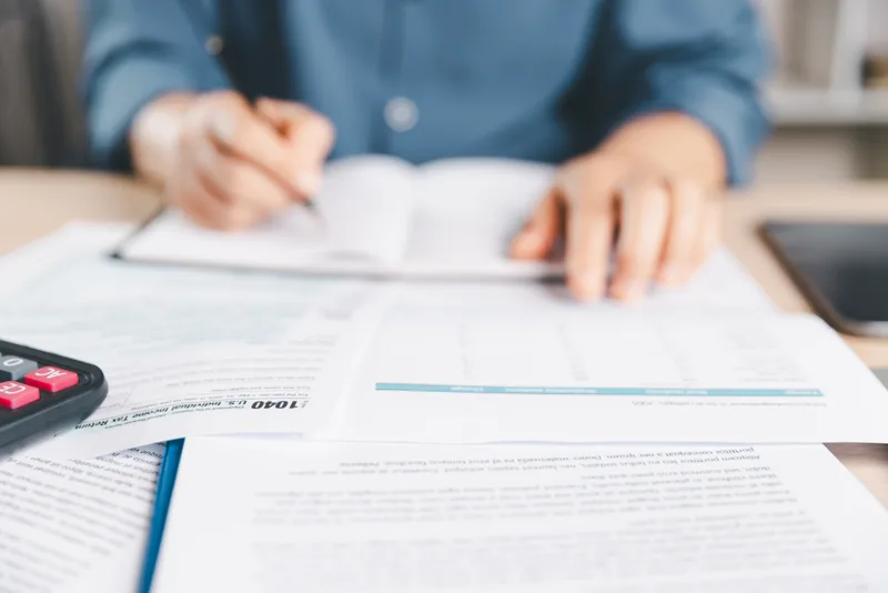 Close view of a preparer's hand signing a Form 1040 at a desk, a calculator at the edge of the paperwork