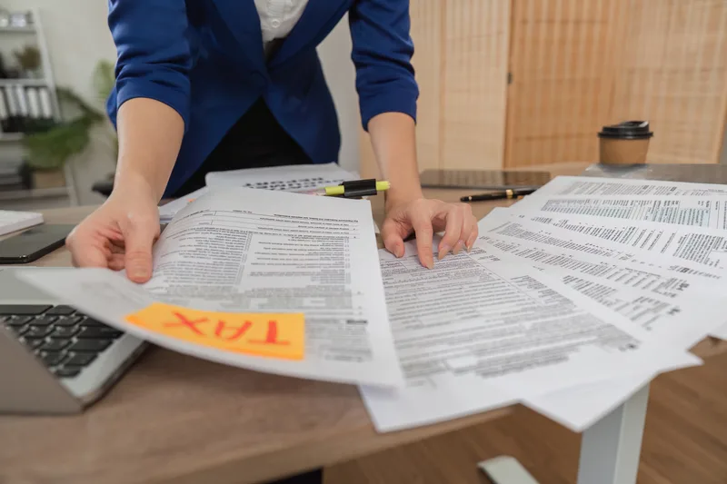 A preparer standing at a desk sorting printed tax forms, with an orange sticky note that says TAX on top of the pile