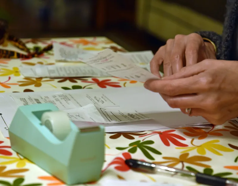 Hands sorting paper receipts on a flowered kitchen tablecloth next to a roll of tape