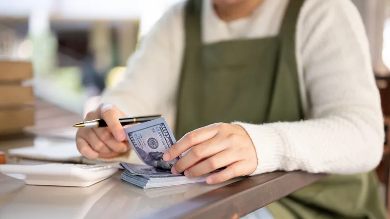 A small shop owner in a green apron counting the day's cash at the counter next to a stack of receipts