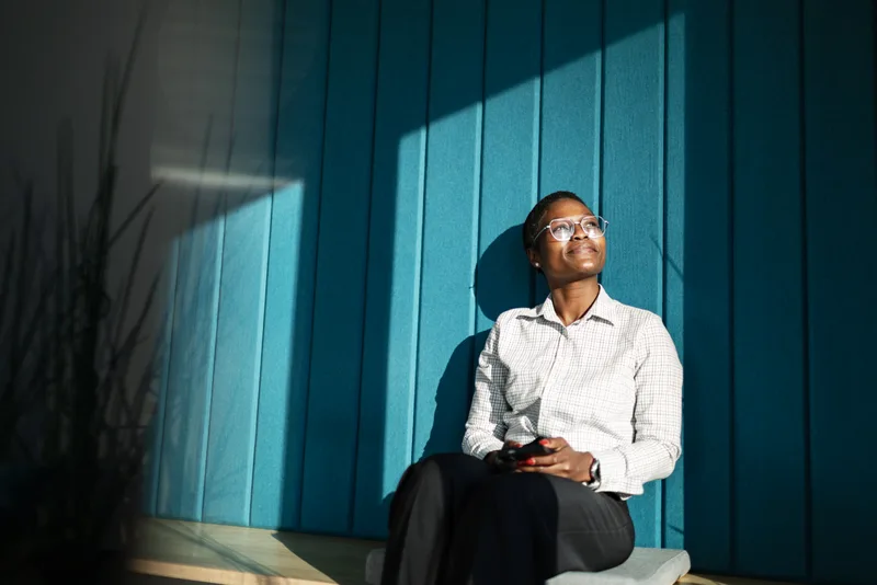 Maya Okafor, Steadwell's founder, seated against a deep blue wall in warm sunlight, looking up thoughtfully