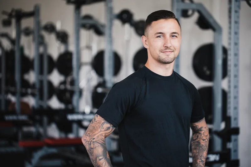Coach Reuben standing on his gym floor in a black t-shirt, tattooed arms relaxed, squat racks behind him
