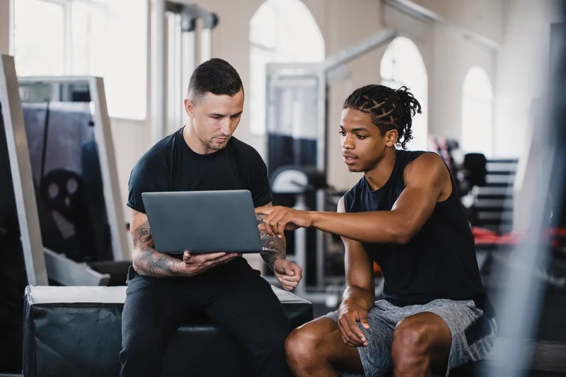 Coach Reuben reviewing a client's training video together on a laptop, sitting on a bench in the gym