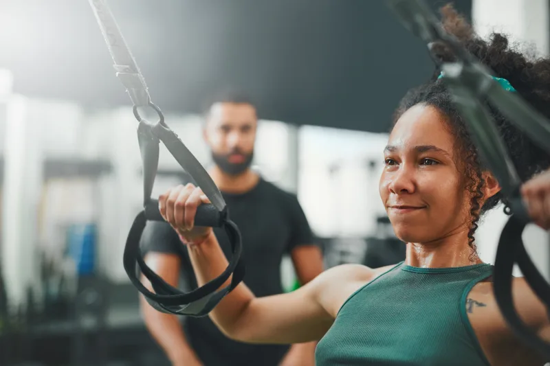 Dana mid-set on suspension trainer straps, focused, with her coach watching from across the gym floor