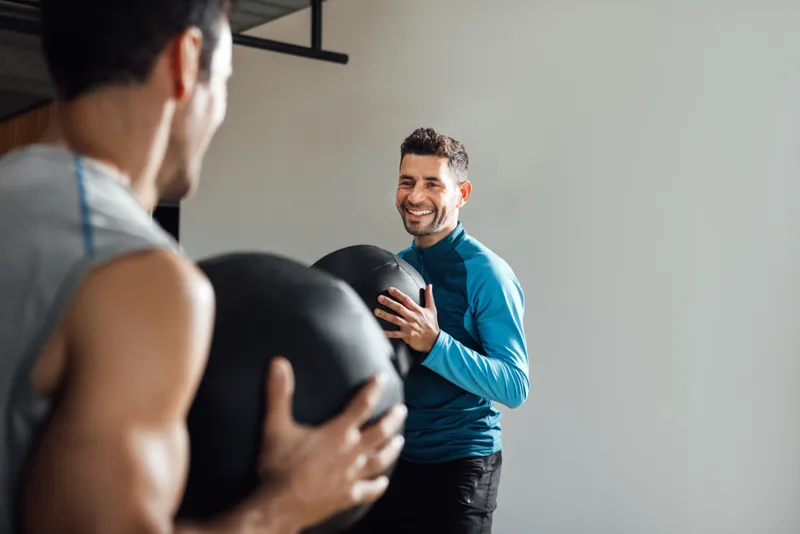 A coach in a training top laughing with a client while handing over a medicine ball between sets