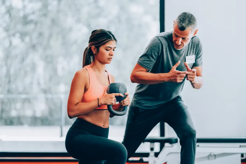 A coach crouched beside a lifter mid-set, cueing her kettlebell position by a bright gym window