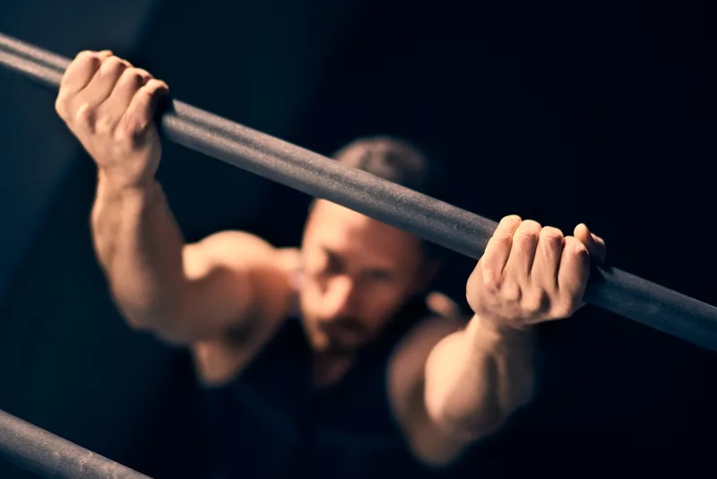Chalked hands gripping a pull-up bar in a dark gym, the lifter's face out of focus below