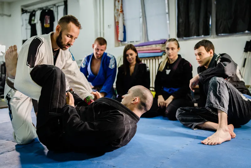 Jiu-jitsu coach in a white gi demonstrating a guard technique on a training partner while the class watches from the mat