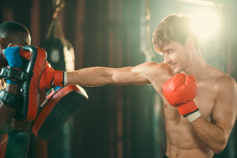 Kickboxer in red gloves extending a straight punch into a coach's curved striking pad in a dim gym